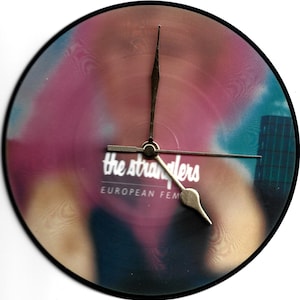 Puede incluir: Un reloj de vinilo negro con una imagen borrosa de una persona y el texto "The Stranglers European Femme Fatale" impreso en la parte delantera.