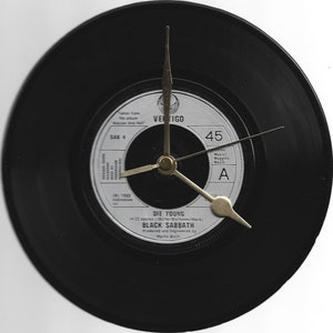 Op de afbeelding: Een ronde klok gemaakt van een zwarte vinylplaat. De wijzerplaat is het label van de plaat, met de tekst "DIE YOUNG" en "BLACK SABBATH". Goudkleurige wijzers geven de tijd aan. Een uniek decoratie-artikel.