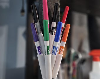 May include: A selection of brightly coloured dry-erase markers in a white holder. The markers are in shades of pink, green, blue, orange, and black. The holder is attached to a dark grey eraser and a black base.