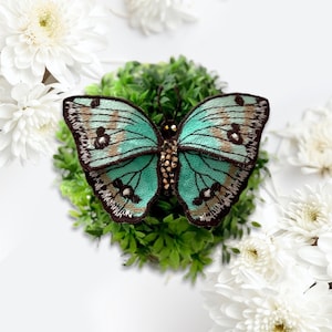 May include: A teal blue butterfly brooch with brown and gold accents. The butterfly is embroidered with rhinestones and has a detailed design.