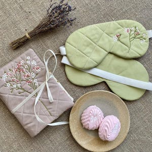 Puede incluir: Dos máscaras para ojos verdes con costuras blancas y bordados florales. Las máscaras están sobre una superficie de tela marrón claro. Un cuaderno acolchado rosa con bordados florales también está sobre la superficie. Un plato pequeño con dos malvaviscos rosas está en la esquina inferior derecha.
