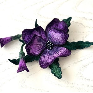 Puede incluir: Un broche de flor bordada de color morado con hojas verdes. La flor tiene un centro morado oscuro con un strass negro.