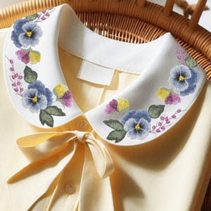Peut inclure: Un col Claudine blanc orné de broderies de pensées bleues, violettes et jaunes. Le col est fixé à un chemisier jaune pâle avec un nœud assorti. Les pensées sont entourées de feuilles vertes et de tiges roses délicates.