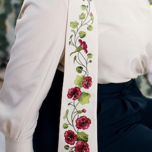 Könnte beinhalten: Cremefarbener Schal mit einem gestickten Blumenmuster. Die Stickerei zeigt rote Blüten, grüne Blätter und zarte Ranken. Der Schal ist über einer cremefarbenen Bluse drapiert, kombiniert mit dunkelgrüner Hose.