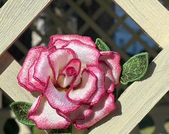May include: A pink and white embroidered rose with green leaves, attached to a white wooden lattice.