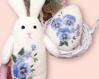 May include: White plush bunny and egg with embroidered purple and blue flowers. The bunny has a pink nose and black eyes. The egg is in a white wicker basket.