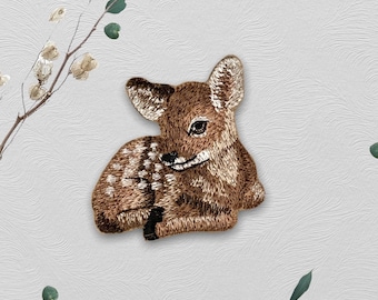 以下が含まれることがあります： 茶色と白の刺繍でできた斑点模様の鹿のアップリケ。鹿は横向きに寝そべっている。