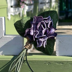 Può includere: Un fiore ricamato viola con foglie verdi e uno stelo di spago marrone. Il fiore è su una recinzione di legno bianca e verde.