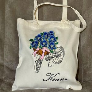 Puede incluir: Una bolsa de lona blanca con un diseño bordado de una bicicleta con una cesta llena de flores azules. El nombre "Kranx" está bordado debajo de la bicicleta.