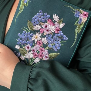 May include: A dark green fabric clutch with a floral embroidery design. The embroidery features a bouquet of pink, white, and blue flowers.