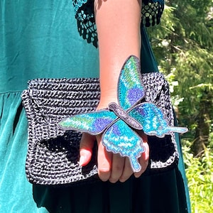 Pode incluir: Uma clutch preta tecida com uma grande aplicação de borboleta azul iridescente. A clutch é segurada por uma mão que veste um vestido verde.