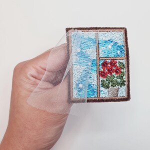 Puede incluir: Parche bordado que representa una ventana con vista a un cielo nocturno estrellado y una planta en maceta con flores rojas. El parche es marrón con una superposición de tela de tul blanco.
