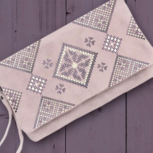 Puede incluir: Un bolso de mano de color rosa claro con un patrón geométrico bordado en hilo blanco y morado. El patrón presenta una forma de diamante en el centro y triángulos más pequeños en los lados.