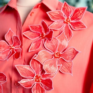 May include: A coral pink button-down shirt with five large red embroidered flowers on the front. The flowers have a white outline and are layered on top of each other.