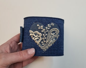 Puede incluir: Un brazalete de tela azul oscuro con un diseño de corazón bordado en oro. El corazón presenta un patrón floral.