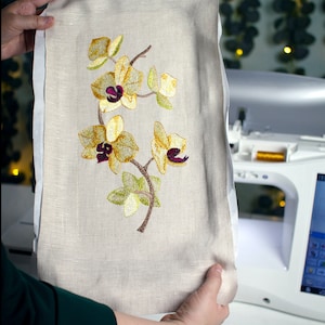 Puede incluir: Una tela beige con un diseño bordado de orquídeas amarillas con centros morados. La tela está siendo sostenida por las manos de una persona.