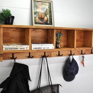 May include: A wall-mounted wooden storage unit featuring five open cubby compartments and seven black metal double hooks. The unit is crafted from light brown wood with a visible grain and includes books, a small plant, and a framed picture.
