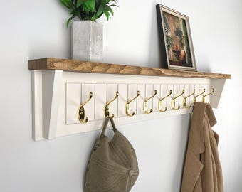 May include: A white wooden wall shelf with a natural wood top and gold hooks. A framed picture and a potted plant sit on the shelf. A tan hat and coat hang from the hooks.
