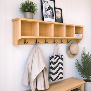 May include: A light-coloured wooden wall-mounted storage unit featuring five open cubby compartments on top and six black metal hooks underneath. The unit is mounted on a white panelled wall and holds a beige jacket, a black and white wavy patterned tote bag, and a straw hat.