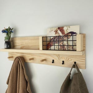 Può includere: Una mensola da parete in legno chiaro con un ripiano, un cestino in filo metallico e ganci. Un cappotto marrone e una borsa verde sono appesi ai ganci. Un piccolo vaso con fiori blu è posto sulla mensola.