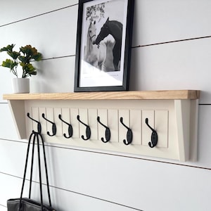 Op de afbeelding: Een aan de muur gemonteerd rek met een licht houten bovenkant en een crèmekleurige basis met acht zwarte haken. Op de plank staat een zwart-witfoto van paarden in een lijst. Een zwarte draagtas hangt aan een van de haken.
