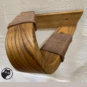 Pode incluir: Prateleira de madeira para gatos com design curvo. A prateleira é feita de madeira castanha clara com grão visível e é enrolada com corda castanha nas juntas. Montada numa parede de tijolo branco.