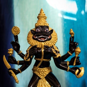 May include: Black and gold statue of a Hindu deity with multiple arms holding various objects. The deity has a fierce expression and is standing on a black base.