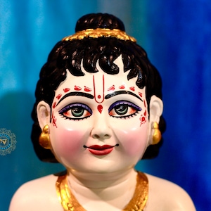 May include: A close-up of a small, white figurine with black hair, a gold crown, and red and white painted details on the face. The figurine is smiling and has a gold necklace.