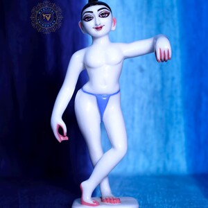 May include: A white figurine of a young person with a blue thong, standing on a white base. The figurine has black hair, painted facial features, and red accents on the hands and feet. The background is a gradient of blue.
