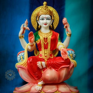 May include: A detailed statue of a deity, seated on a pink lotus flower. The figure wears a red sari, green blouse, and gold jewelry, including a crown and halo. The background is a gradient of blue and teal.