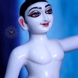 May include: A white marble statue of a person with black hair and painted facial features, including pink lips and blue eyes. The figure is wearing a blue garment around the waist and has one arm extended. The background is a gradient of blue.