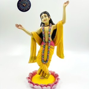 May include: A figurine of a Hindu deity standing on a lotus flower base. The deity is wearing a yellow robe and a garland of flowers. The figurine is made of painted resin.