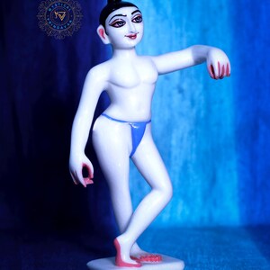 May include: A white figurine of a person in a dancing pose, with black hair and facial features. The figurine wears a blue thong and has pink accents on the hands and feet. It stands on a white rectangular base against a blue backdrop.