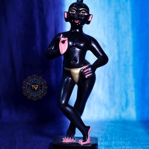 May include: A black figurine of a person with pink accents on the ears, hands, and feet, and a gold-colored loincloth. The figurine is standing on a black base. The background is a gradient of blue.