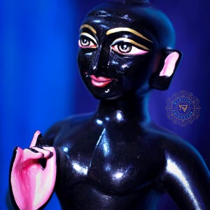 May include: A glossy black figurine of a person with gold accents on the eyes and a pink hand gesture. The figure has a small topknot and pink accents on the ears and lips. The background is a gradient of blue.