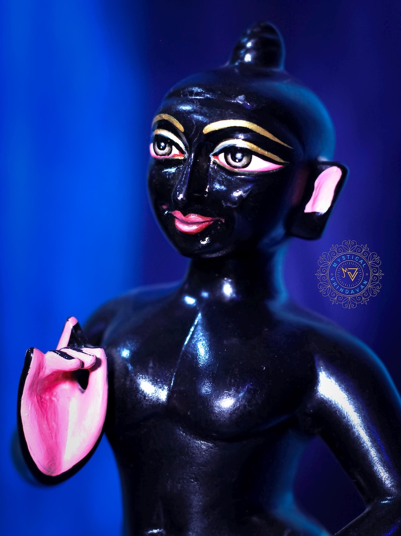 May include: A glossy black figurine of a person with gold accents on the eyes and a pink hand gesture. The figure has a small topknot and pink accents on the ears and lips. The background is a gradient of blue.
