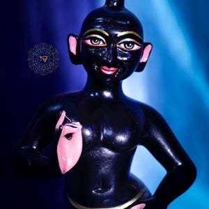 May include: A glossy black figurine of a person with gold accents on the eyes and a gold loincloth. The ears and hands are painted pink. The figure is posed with one hand raised and the other resting on the hip.