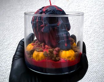 Op de afbeelding: Een zwarte en rode schedelkaars met een rode wasdruppel, omringd door kleine gele pompoenkaarsen in een glazen pot.