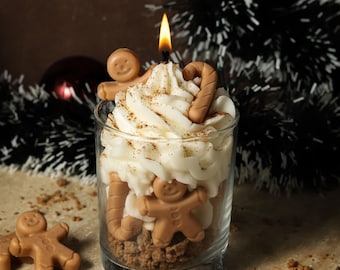 May include: A clear glass jar filled with a gingerbread-themed candle. The candle is topped with white frosting and decorated with gingerbread men and candy canes. The candle is lit and the flame is visible.