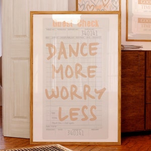 May include: A framed print with a white background and black grid lines. The text "Dance More Worry Less" is written in orange on the print. The print is framed in a light wood frame.