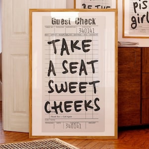 Puede incluir: Tres impresiones enmarcadas con texto humorístico sobre un fondo blanco. La impresión más grande dice "TAKE A SEAT SWEET CHEEKS". Las otras impresiones dicen "THIS MUST BE THE" y "Go piss girl". Los marcos son de color marrón claro.