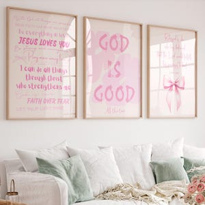 May include: Three framed wall art prints featuring pink text and graphics on a light background. The center print displays the words GOD IS GOOD in large pink letters. The side prints contain various religious phrases and a pink ribbon illustration.