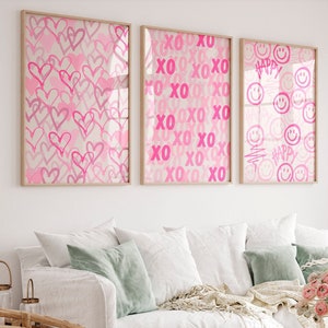 Peut inclure: Trois cadres muraux rectangulaires. L'affiche de gauche présente un motif de cœurs roses dessinés à la main. L'affiche centrale affiche un motif répétitif XO rose. L'affiche de droite montre des visages souriants roses et le mot HAPPY.