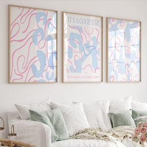 May include: Three framed wall art prints featuring abstract pink and light blue organic shapes on a white background. The center print includes the text IT'S A GOOD DAY TO HAVE A GOOD DAY in a simple sans-serif font, displayed in light blue and pink.