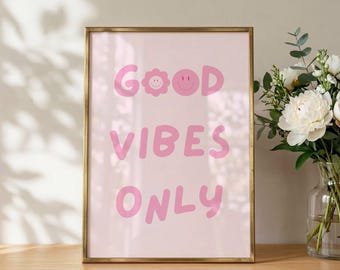May include: A rectangular wall art print in a thin gold frame featuring the text GOOD VIBES ONLY in pink bubble letters. The letter O in GOOD is replaced by a pink flower shape and a pink smiley face. The background of the print is light pink.
