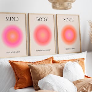 May include: Three framed art prints with the words "MIND", "BODY", and "SOUL" above a vibrant pink and orange gradient circle. The prints also include the phrases "FREE YOUR MIND", "NOURISH YOUR BODY", and "FEED YOUR SOUL". The prints are displayed above pillows.