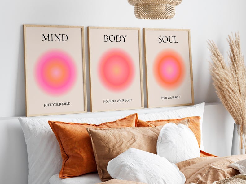 May include: Three framed art prints with the words "MIND", "BODY", and "SOUL" above a vibrant pink and orange gradient circle. The prints also include the phrases "FREE YOUR MIND", "NOURISH YOUR BODY", and "FEED YOUR SOUL". The prints are displayed above pillows.