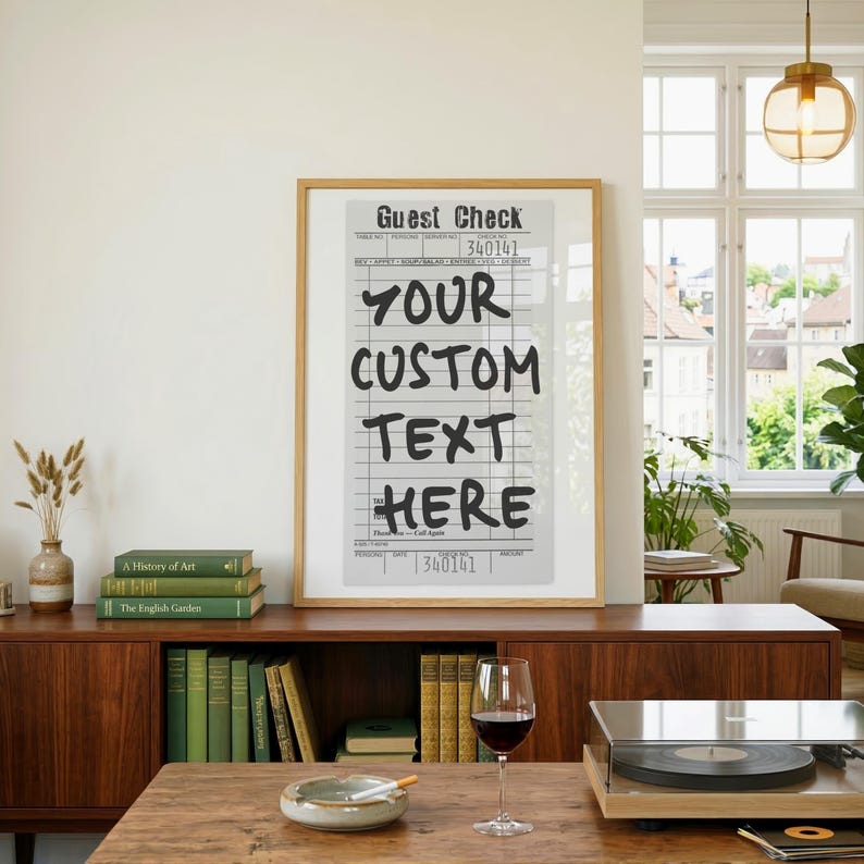 May include: A framed wall art print featuring a vintage-style guest check design with the text "GUEST CHECK" at the top and "YOUR CUSTOM TEXT HERE" in large black lettering, displayed in a light wood frame on a wooden sideboard.