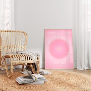May include: A pink and white poster with the text "Angel No. 444 Protection" and a pink circle in the center. The poster is hanging on a white wall next to a wicker chair and a jute rug.