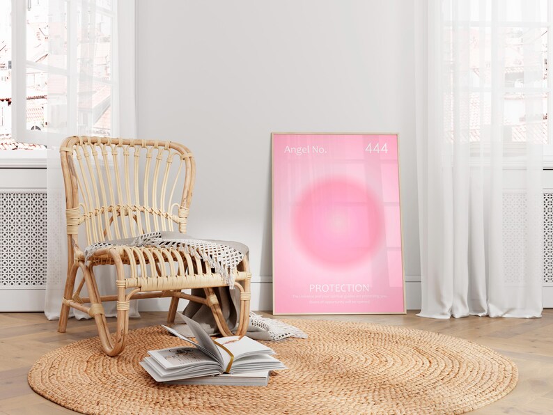 May include: A pink and white poster with the text "Angel No. 444 Protection" and a pink circle in the center. The poster is hanging on a white wall next to a wicker chair and a jute rug.